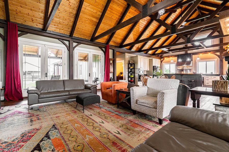 Photo of great room and kitchen of log home with exposed light wood ceiling and dark a-frame trusses. White framed glass windows lead to an porch