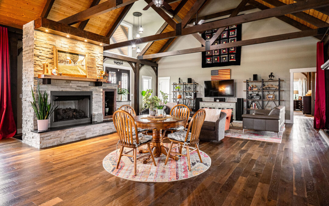 Photo of great room with light wood ceiling, dark a-frame trusses and dark wood floors. a large stone fireplace is on an exterior wall