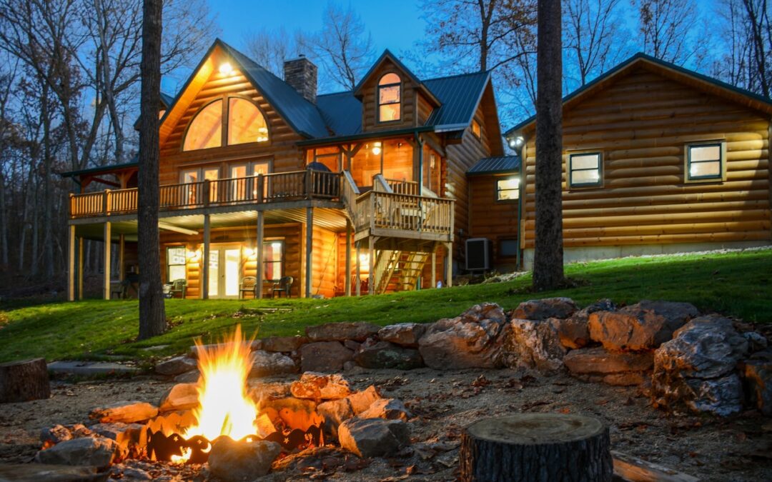 Photo of rear of log home on heavily treed lot with rustic steps leading away from the home. The photo was taken at dusk with a bon fire in the backyard.