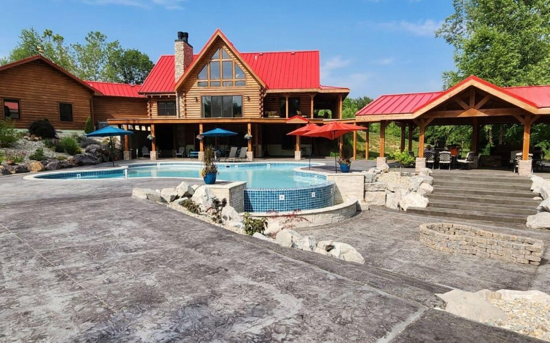 Photo of log home estate with bright red roof on the home and the pool pavilion. the photo is taken from the patio and pool deck showing the rear of the home with exposed wood, covered upper level porches and garage attached to the home by a breezeway.