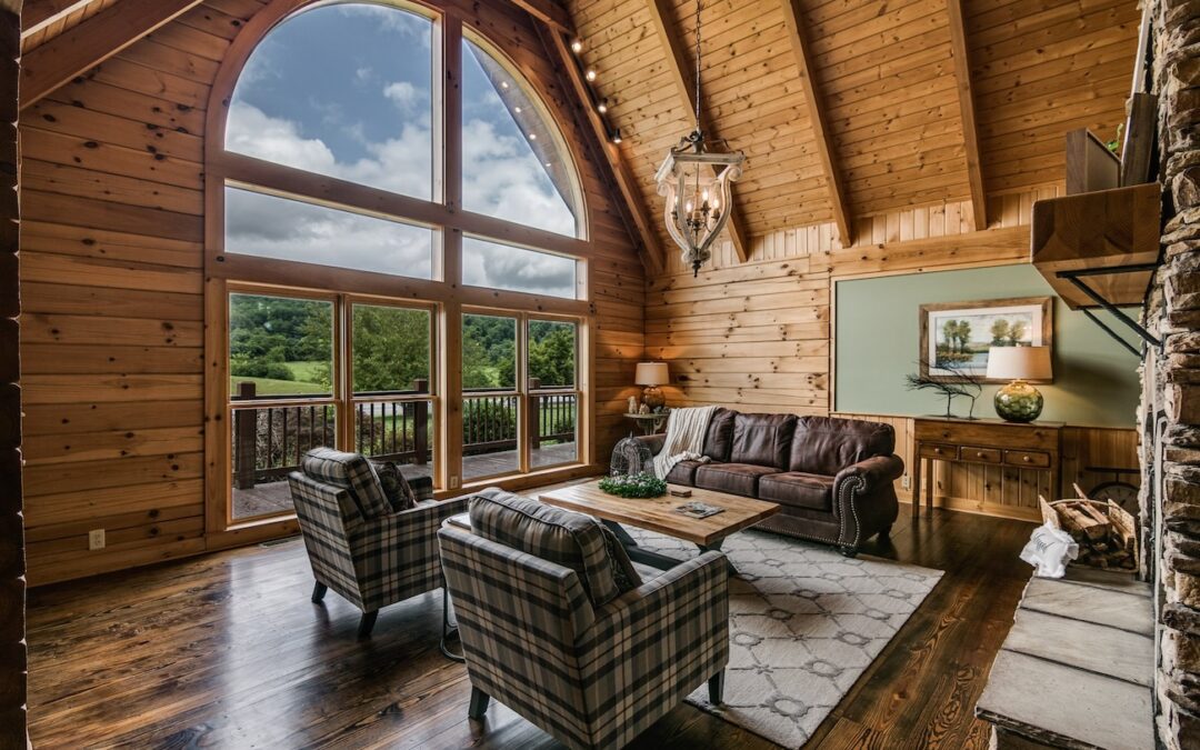 Photo of great room with exposed wood ceiling and high truss supports. Exterior wall features paladium style geometric upper windows over two sets of glass doors leading to deck