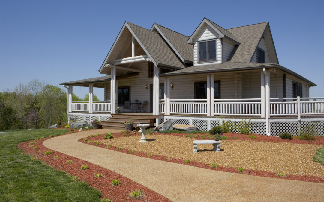 Photo of log home with whitewashed wood exterior walls and white railings on the wrap around porch and deck system. An a frame dormer over the front entrance and an upper dormer on the second floor of the home create a cottage appearance