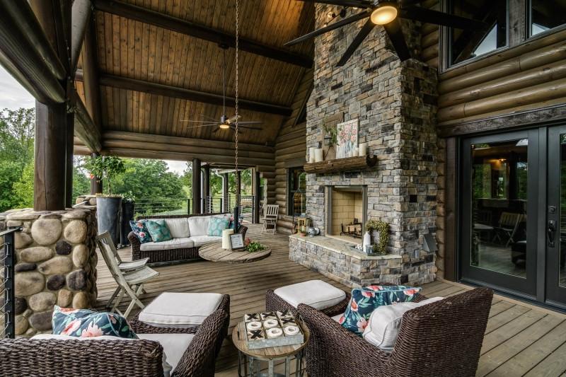 Photo of outdoor living space with stone fireplace room for comfy couches and chairs, ceiling is round logs and heavy wood support beams. Lighted ceiling fans are installed in the ceiling.