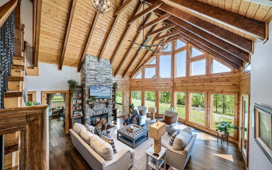 Photo of great room taken from stairs leading to loft area of log home. The roof is exposed wood with darker wood truss system. the exterior wall is matching exposed wood with upper wall geometric windows and entire lower wall giant windows. A stone fireplace bisects the great room creating a "room" feel separating a guest room area and doors leading to outdoor living space