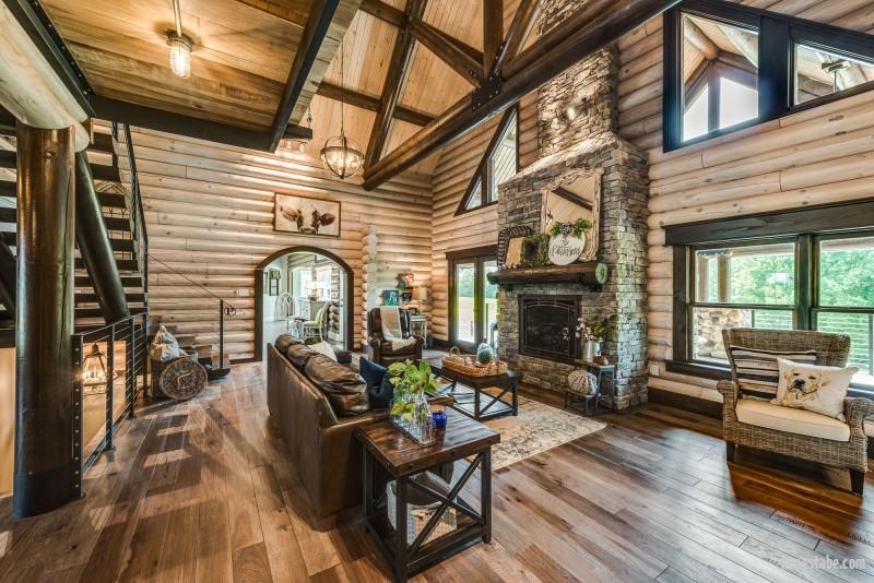 Photo of great room with exposed round logs, soaring stone fireplace soaring ceilings of exposed wood with dark wood a-frame trusses. Upper windows on exterior wall are geometric around fireplace chimney . lower windows are casing along with glass doors leading to porch