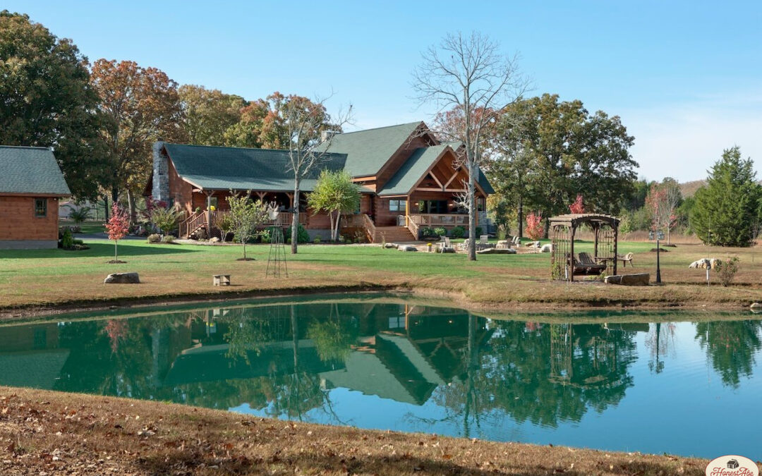 Photo of log home estate home taken from across large pond area. There are matching log outbuildings on the property. The buildings all have green colored roofs