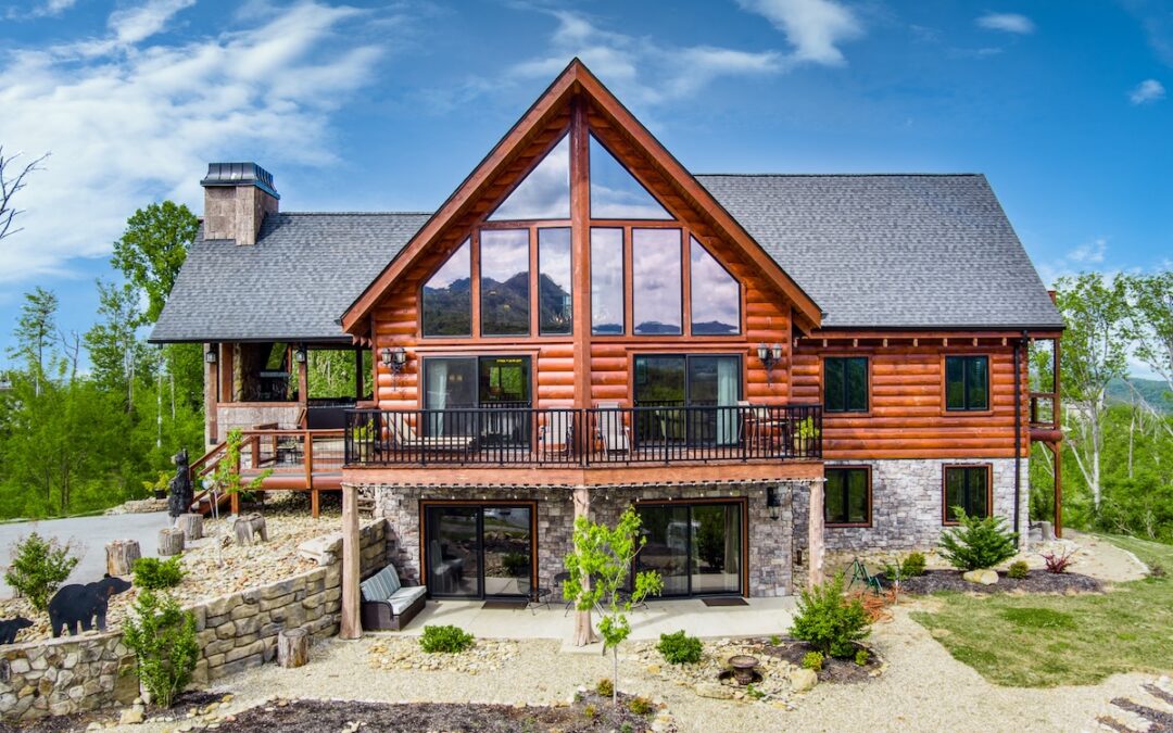 Photo of rear of log home with stone foundation/walk out basement area. Upper levels are round log profile with al large deck off the great room and bedroom. The side entrance of the home has a covered porch living area.